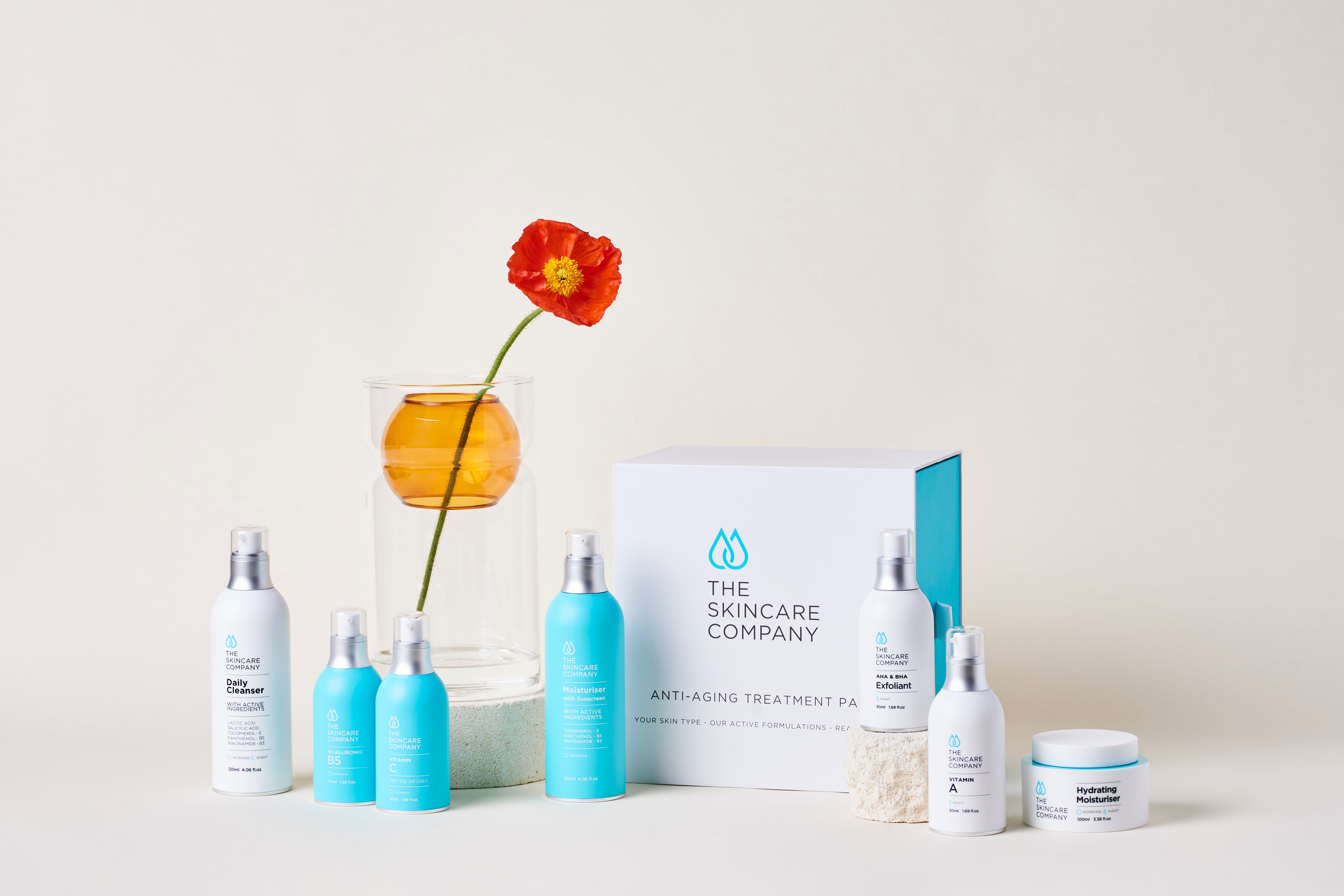 Skincare product set with cleanser, moisturiser, exfoliant, and vitamin serums beside a flower in a vase.